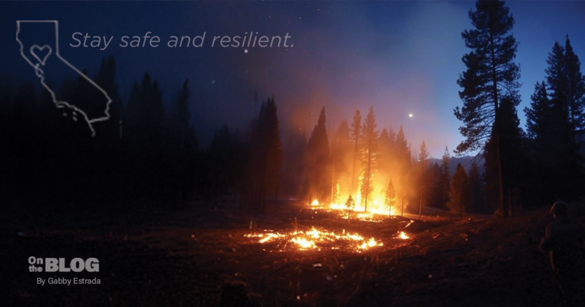 Aerial view of a California wildfire, with flames and smoke rising from a forested area under a hazy sky.